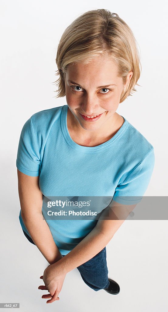 A cute blond caucasian teenage girl wearing a blue teeshirt is looking up at the camera with hands clasped and smiling shyly