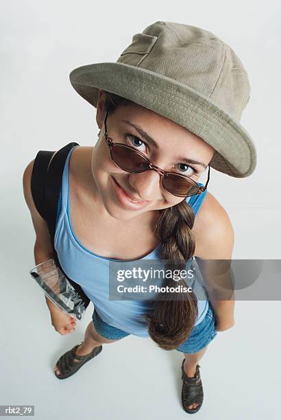 a pretty teenage girl in sunglasses and a hat with a blue tank top and shorts has a long brown braid and a shoulder bag and smiles over her glasses up at the camera - camera bag stock-fotos und bilder