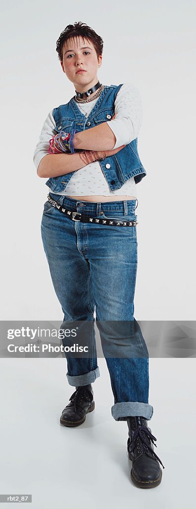A caucasian teenage girl with spiked hair and a studded belt is wearing jean pants and vest as she stands with arms folded and frowns at the camera