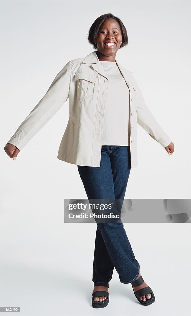 A cute african american teenage girl wearing jeans and a white blazer is standing about to twirl around as she spreads her arms and one foot crosses over the other