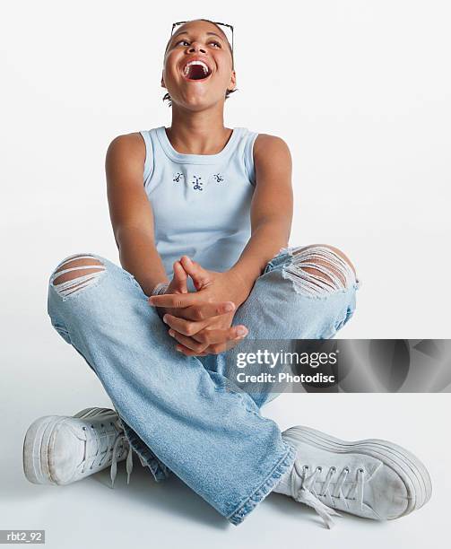 an african american teenage girl wearing jeans with holes in the kness sits crossleg on the floor as she throws her head back and laughs - alleen één tienermeisje stockfoto's en -beelden