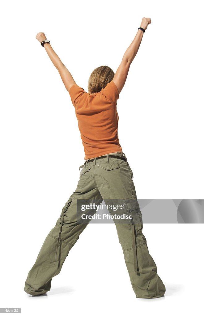 Caucasian blonde female teen in green pants and orange shirt stands backwards with arms in the air