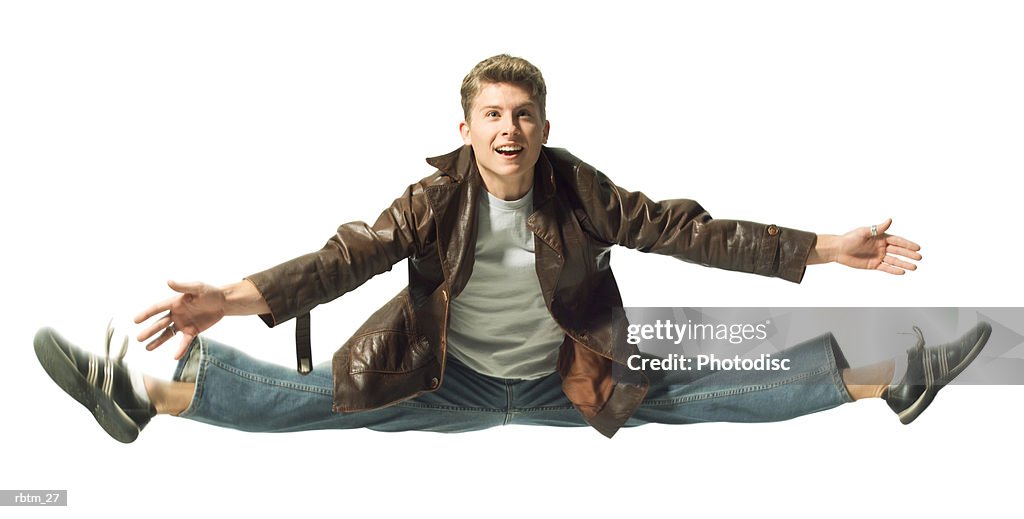 A caucasian male teen in jeans and a leather jacket jumps up and does the splits