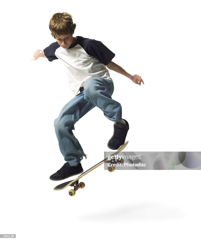 A caucasian male teen in jeans and a t shirt jumps up while riding his skateboard