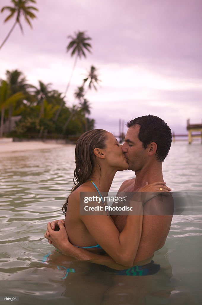 A caucasian couple embrace and kiss in the water as they relax in a tropical locale