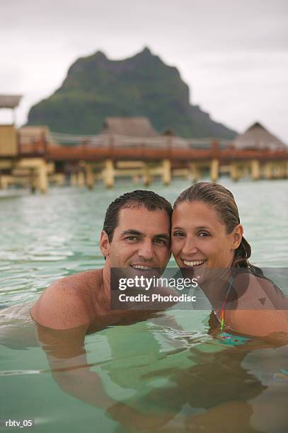 a caucasian couple smile at the camera as they swim in the water at a tropical resort - smile stockfoto's en -beelden