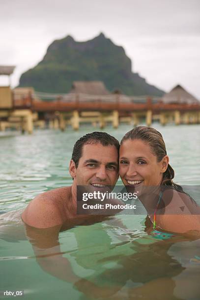 a caucasian couple smile at the camera as they swim in the water at a tropical resort - straw hut stock pictures, royalty-free photos & images