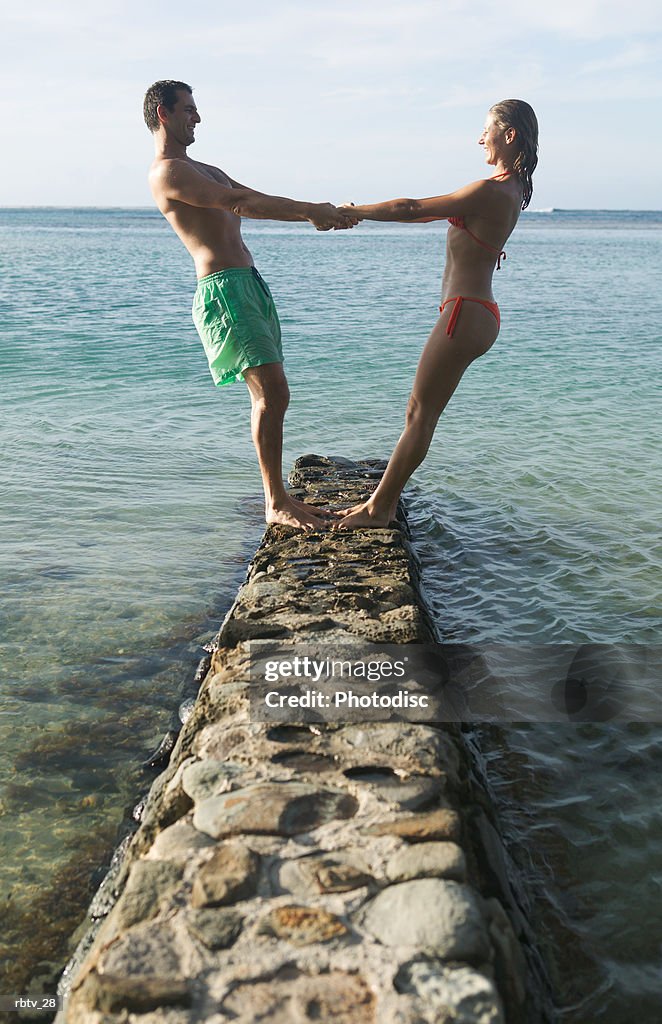 A caucasian couple in swimsuits holds hands and lean back white playing on a stone bridge at the beach