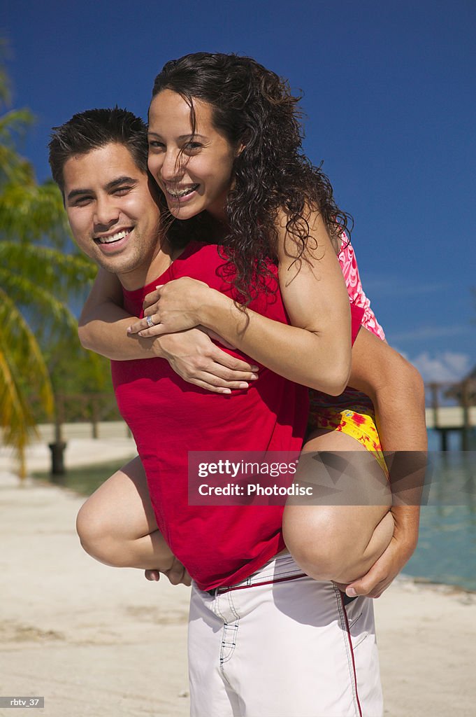 An ethnic woman in a swimsuit plays at the beach with her boyfriend by jumping on his back
