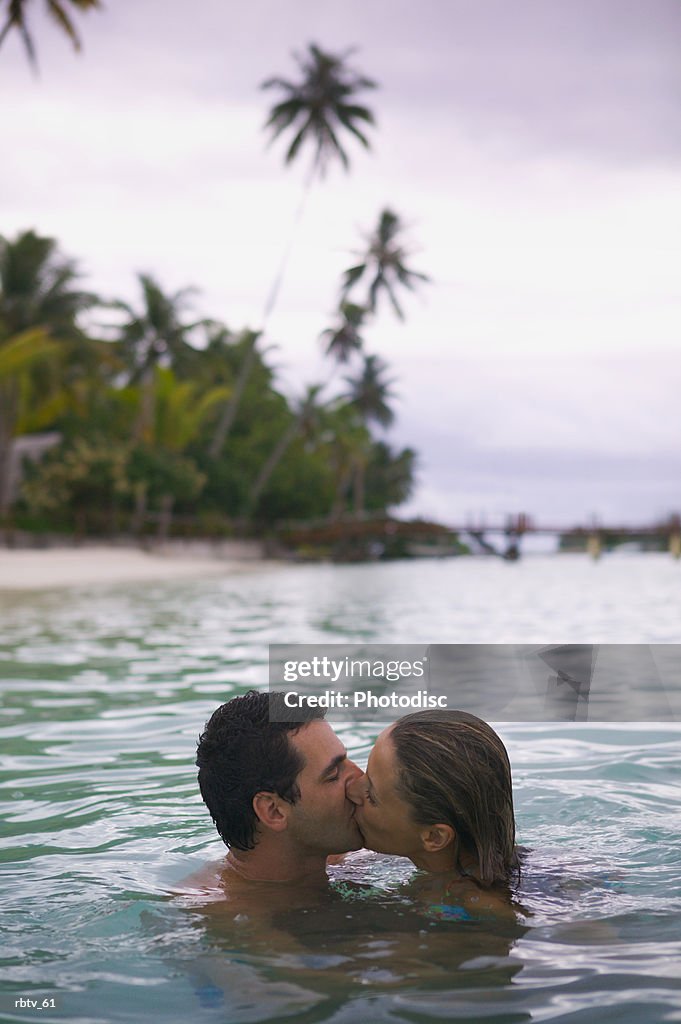 A caucasian couple swim together and kiss in the water while they vacation in the tropics