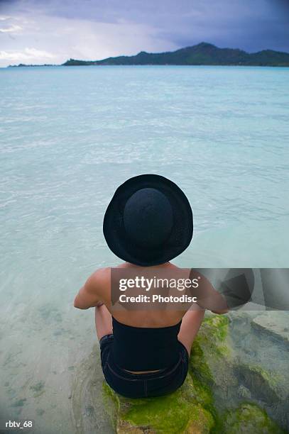 a caucasian woman in a black hat sits in the water on a beach in a tropical setting - gesellschaftsinseln stock-fotos und bilder