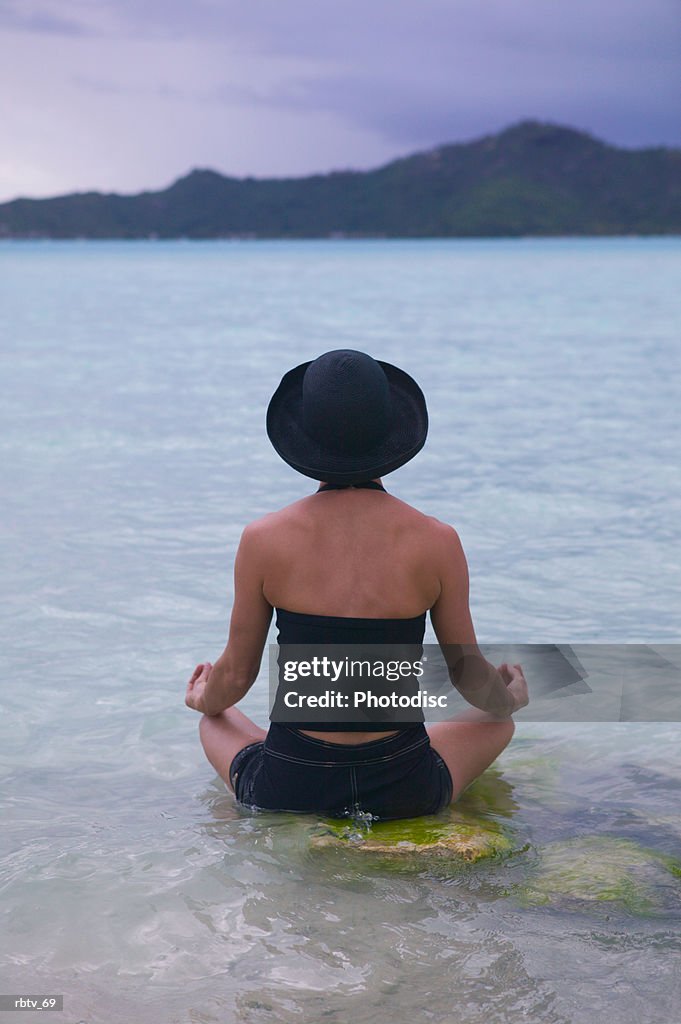A caucasian woman in a black hat meditates in the water on a beach in a tropical setting