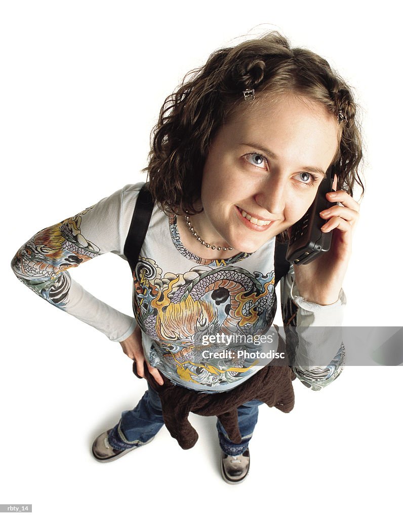 Young caucasian girl student with funky hair wearing a shirt with a dragon painted on the front a sweatshirt around her waist and blue pants carries a backpack and talks on a cellular phone