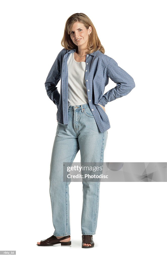 Pretty young caucasian woman with long brown hair wearing a blue shirt sandals and blue jeans stands smiling at the camera with her hands on her hips and smiles