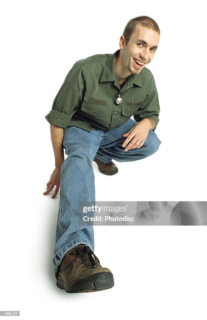 College-age young man with a small beard and short hair wearing a green army shirt blue jeans and brown shoes squats down and sticks one foot out towards the camera and smiles