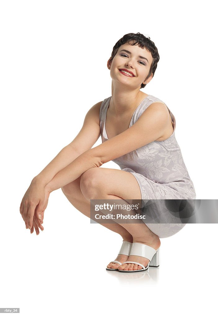Beautiful italian looking young woman with short dark hair beautiful features and red lips wearing a short purple dress and white shoes squats down with arms outstretched in front of her tilts her head and smiles at the camera