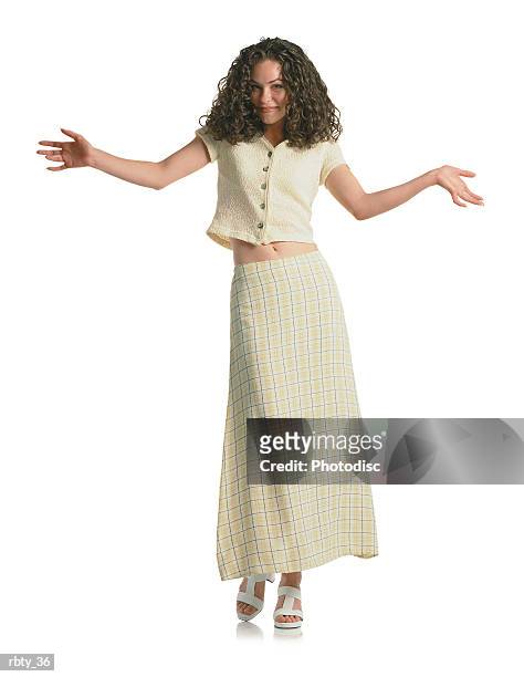 young beautiful brunette teenage girl with curly hair wearing a cream plaid skirt and cream short shirt steps toward the camera and gives a slight smile as she holds her hands and arms out to the side - smile stockfoto's en -beelden