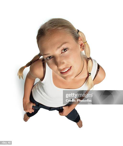 funky teenager with funny blonde hair in pigtails and brown eyes wearing a white tank top and dark pants looks up into the camera with a slight smile as she tilts her head to the side and puts her hands in her pockets - smile stockfoto's en -beelden