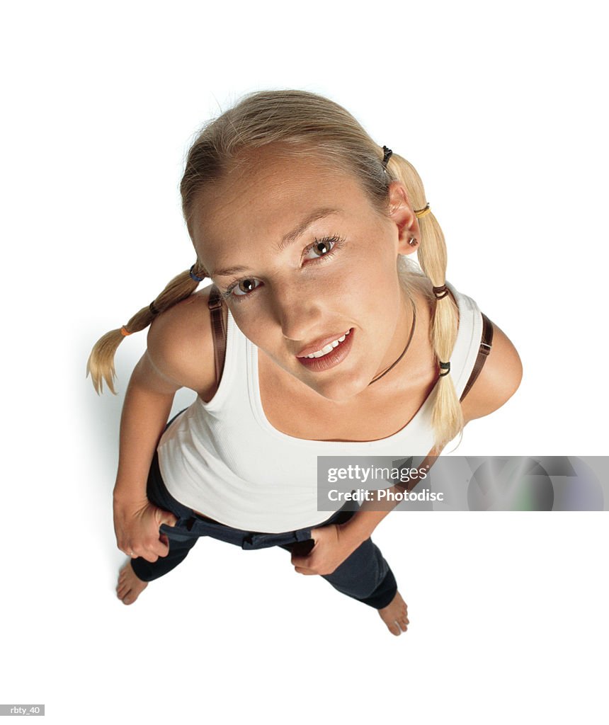 Funky teenager with funny blonde hair in pigtails and brown eyes wearing a white tank top and dark pants looks up into the camera with a slight smile as she tilts her head to the side and puts her hands in her pockets
