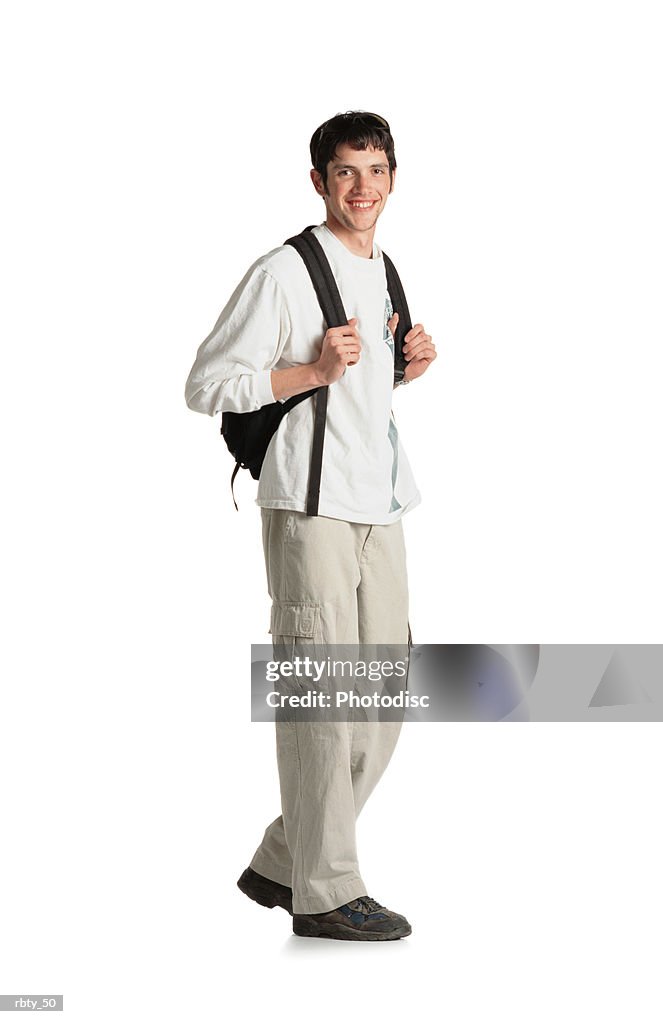 Young college or high school student with dark hair and scruffy beard wearing a long-sleeved white shirt tan pants and brown shoes carries a backpack and walks in front of the camera and smiles