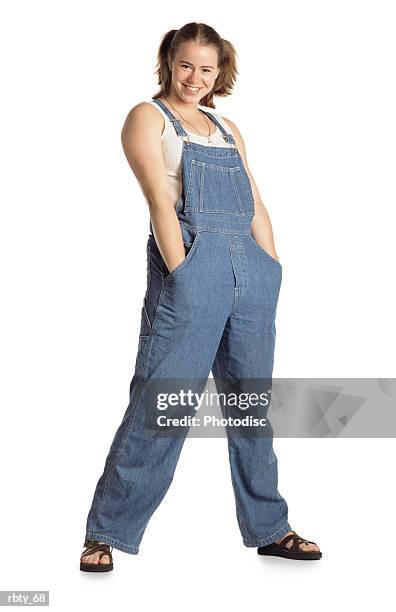 healthy young country girl with pigtails in her short brown hair wearing blue bib overalls over a white tank top with brown sandals and a necklace shoves her hands deep into her pockets with her legs spread apart and smiles at the camera - bib necklace stock-fotos und bilder