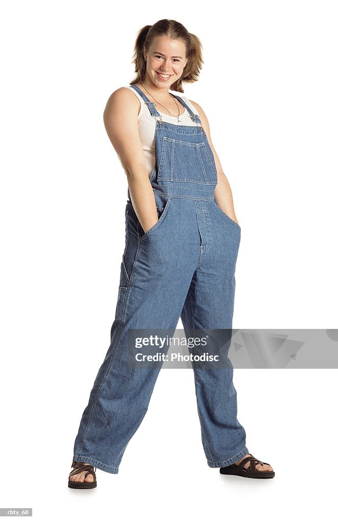 Healthy young country girl with pigtails in her short brown hair wearing blue bib overalls over a white tank top with brown sandals and a necklace shoves her hands deep into her pockets with her legs spread apart and smiles at the camera