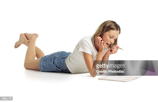 cute teenage caucasian girl with shorter brown straight hair wearing a white shirt and blue denim shorts lays on the ground chewing on a pen while she looks over her homework - white shirt ストックフォトと画像