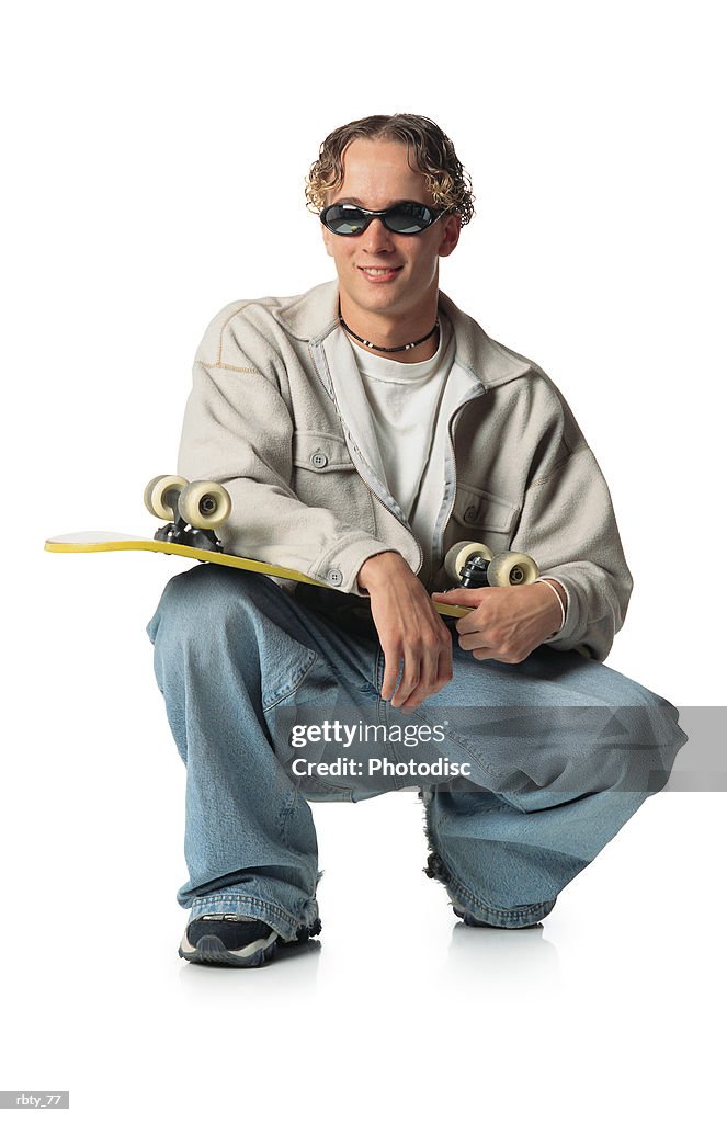 Young caucasian man with brown hair wearing blue denim jeans and a cream long-sleeve jacket over a white shirt and sunglasses and necklace holds a skateboard as he crouches