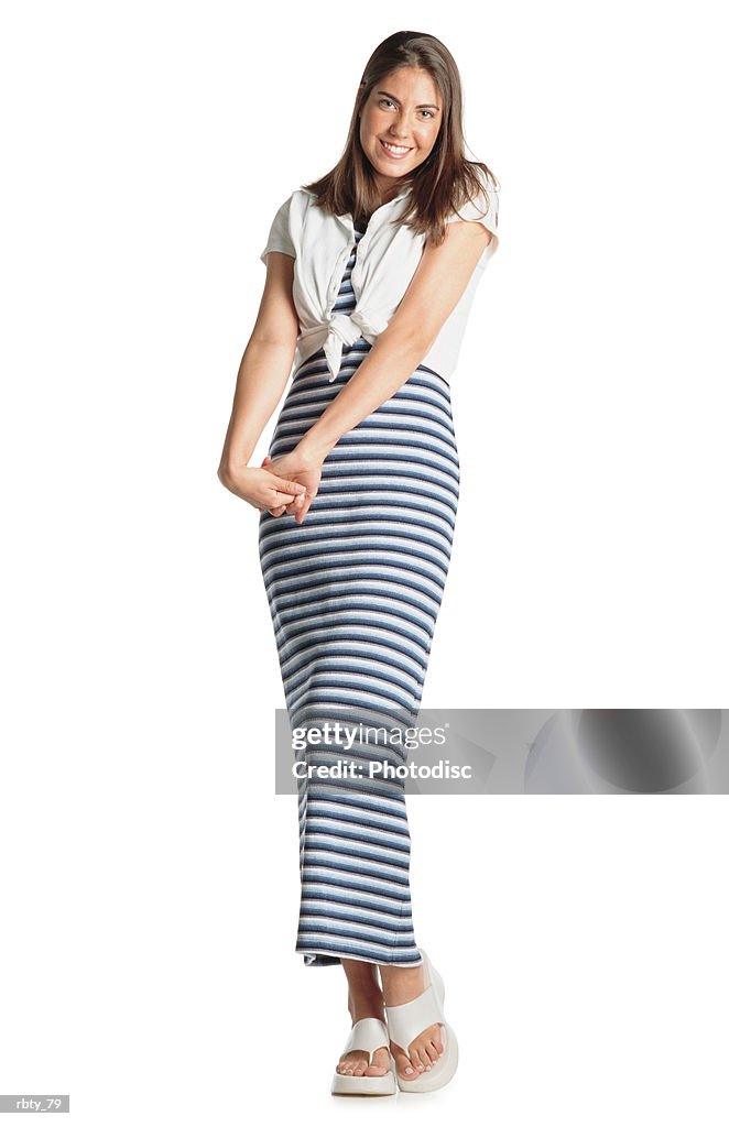 Teenage girl with long brown hair wears a striped dress with a white shirt and white sandles crosses her hands as she smiles at the camera
