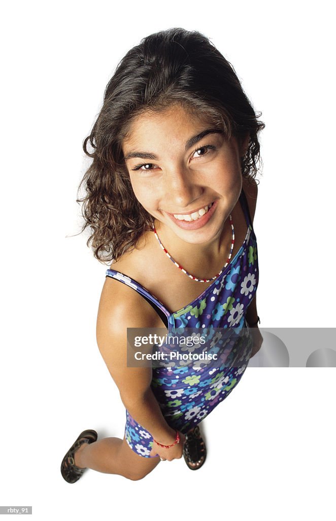 A brunette hispanic teenage girl wearing black sandals and a flower dress with spaghetti straps and necklace smiles as she looks up toward the camera