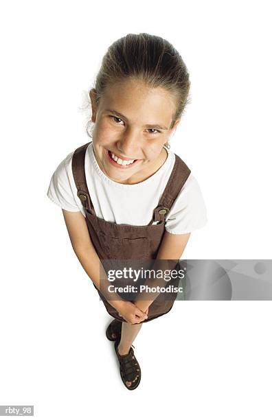 young blond-haired caucasian girl wearing brown sandals and a brown jumper over a short-sleeve white shirt crosses her hands in front of her as she peers upward through brown eyes at the camera and smiles - white shirt ストックフォトと画像