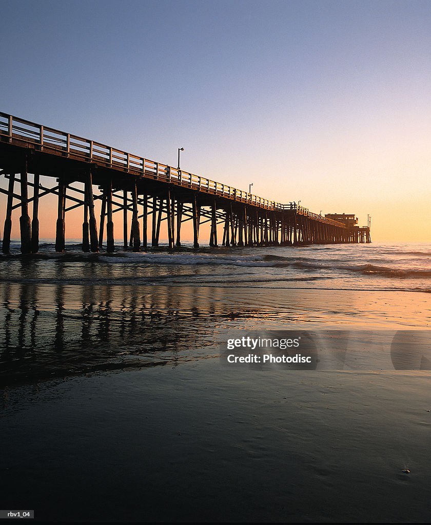 A bridge rises above the ocean waves as the sun sets over the horizon under a blue sky
