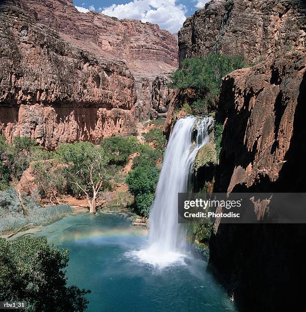 a waterfall falls into blue-green water amongst green trees and jagged mountain cliffs under a blue sky with clouds - plant condition stockfoto's en -beelden