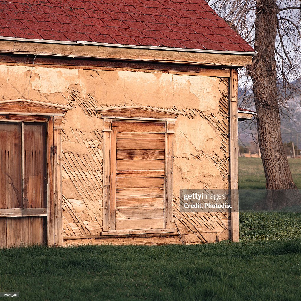 An abandoned house with red shingles and a boarded up doorway and window sits among green grass and trees with the mountains in the distance