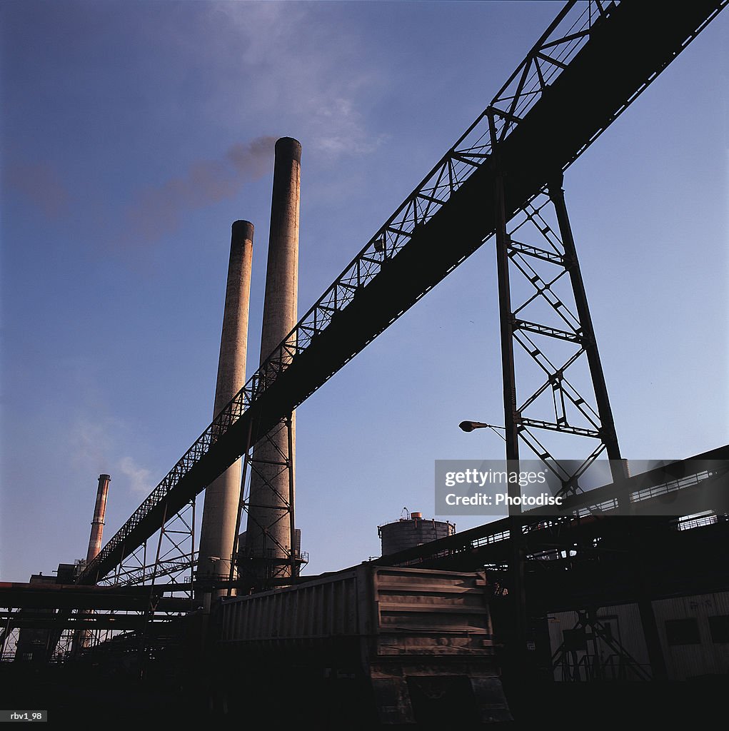 Smoke stacks and other pipes stand under a clear blue sky as smoke leaves the stacks