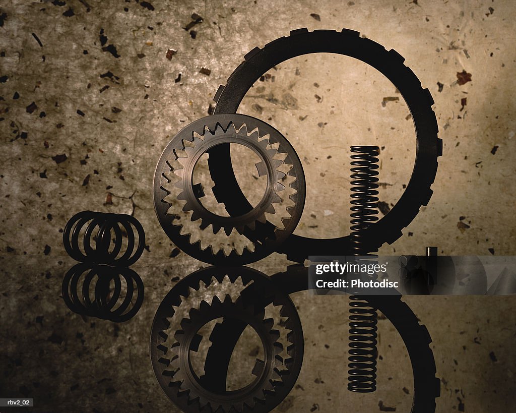 Several gears and a spring stand on a reflective surface with a speckled background