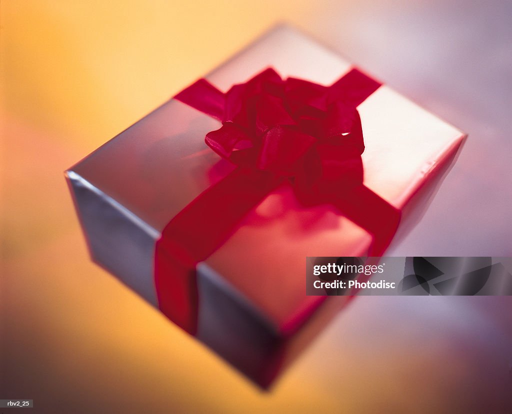 A silver and red bow wrapped gift sits on  a yellow and silver surface as light reflects off it
