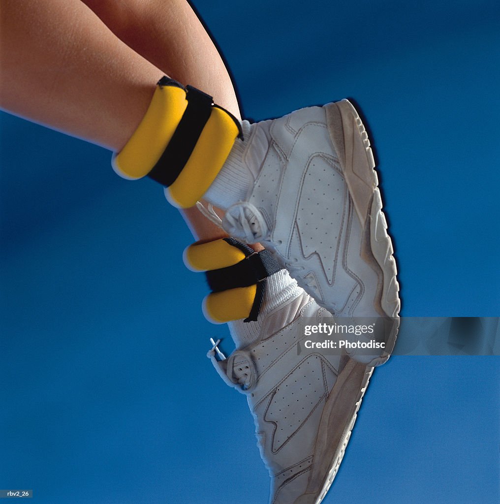 A pair of white shoes and yellow ankle weights rest on legs in front of a blue background