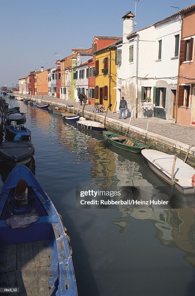 Painted houses and boats line the edge of a reflective canal in Europe under a clear blue sky