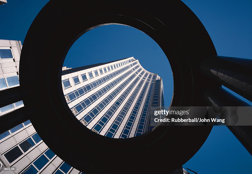Looking up through a large black circle to see a curved building with large black windows reaching toward the clear blue sky