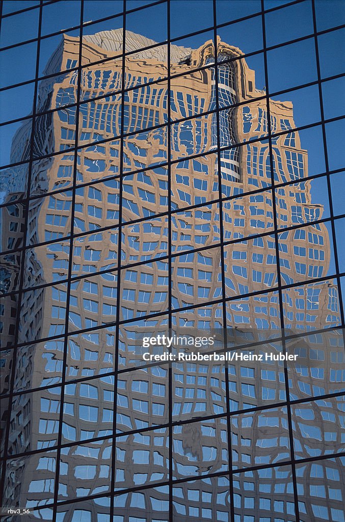 A large windowed beige building is reflected in the glass windows of another skyscraper