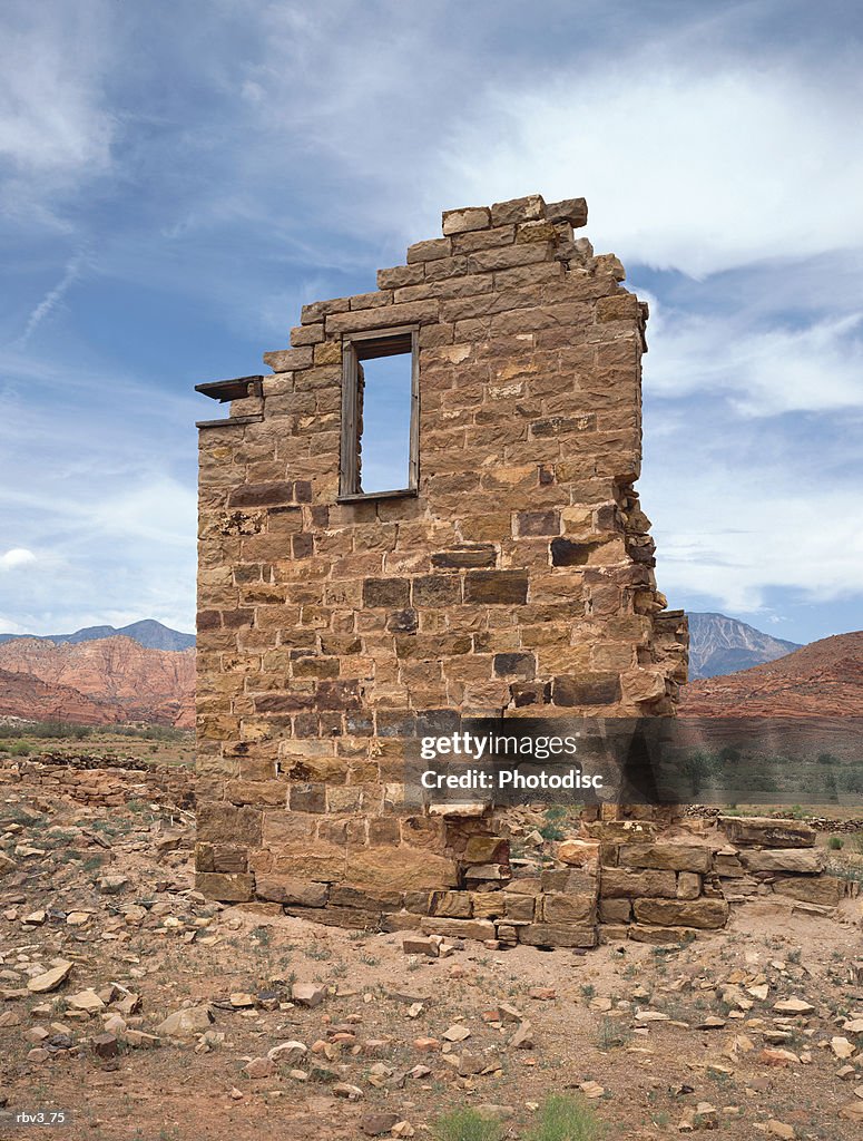 Part of an adobe brick wall with a window opening stands amongst rocks and green plants under a blue sky with white clouds