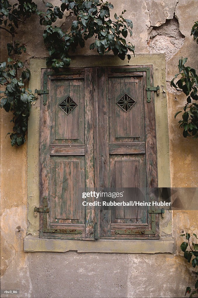 Wooden shutters cover a window in a stone wall with plants hanging from above