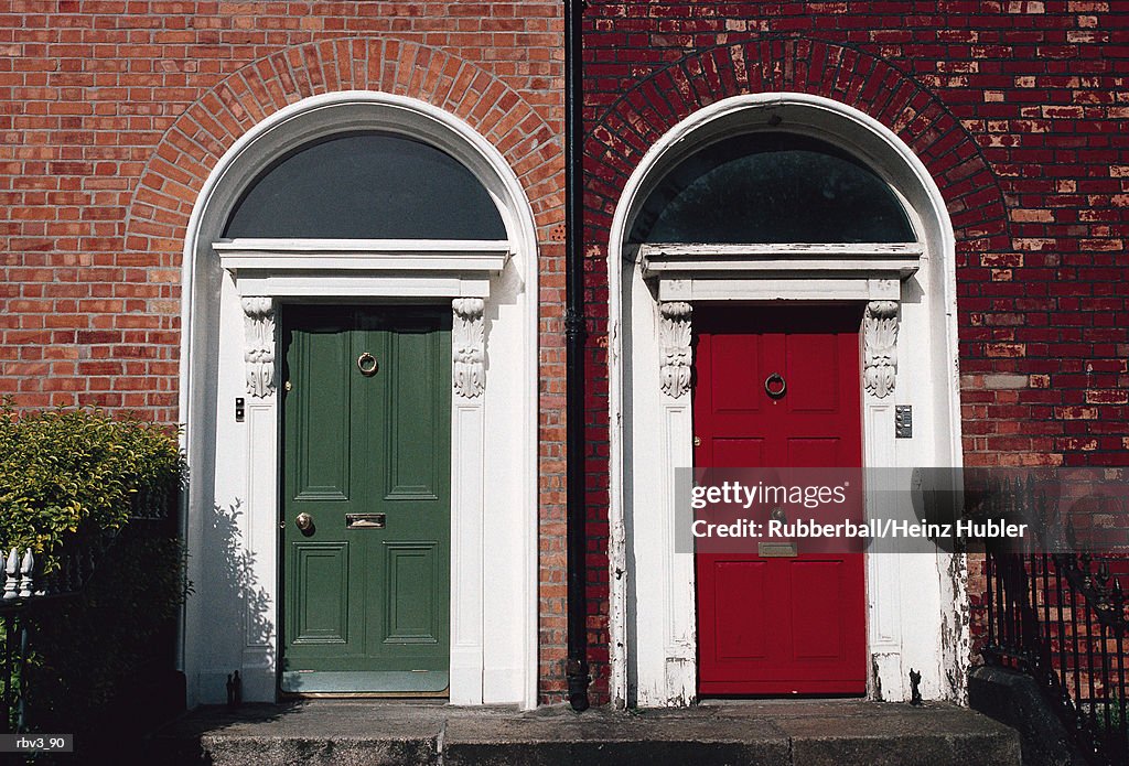 A red and green door from different houses with white frame stand next to each other with railings and green bushes present