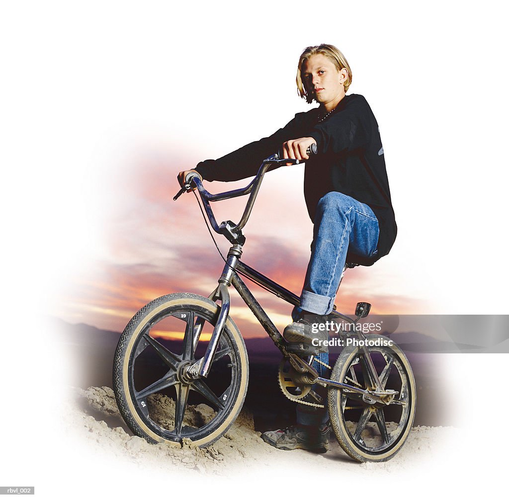 A caucasian boy in a black shirt and blue jeans is sitting on a freestyle bicycle with the sun setting behind him