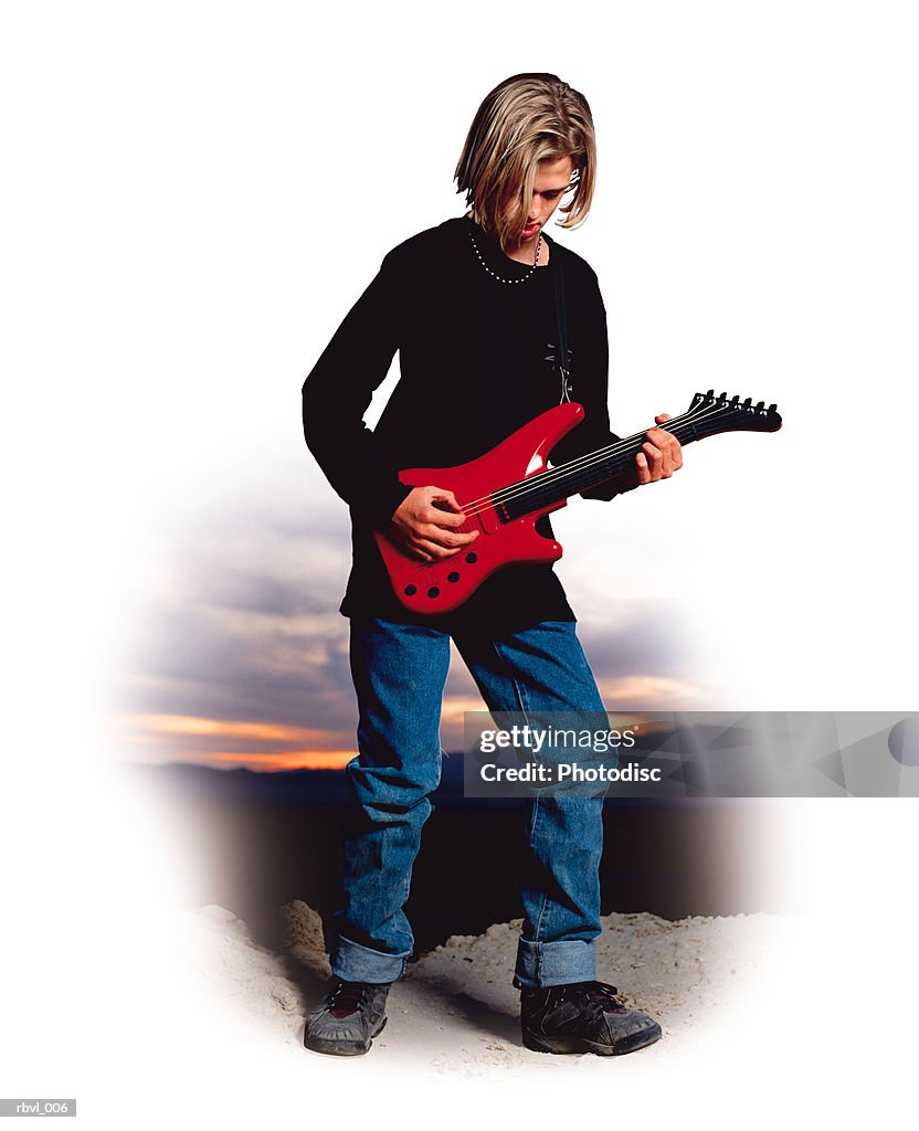 A male caucasian youth with long blond hair wearing a black shirt and blue jeans is standing outside playing an electric guitar