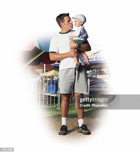 a young caucasian father in gray shorts and a white t-shirt is at a fair holding his baby son who is wearing a white baseball cap - genderblend stock-fotos und bilder