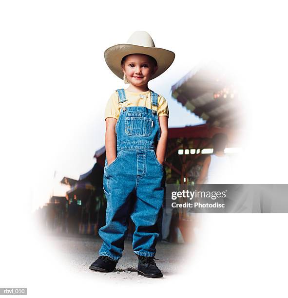 a little caucasian girl in blue overalls and a yellow shirt and a white cowboy hat is standing at a county fair - county_fair stock-fotos und bilder