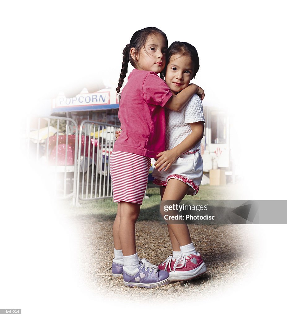 Two little hispanic girls at a county fair are wearing pink and white shorts and t-shirts as they hug each other