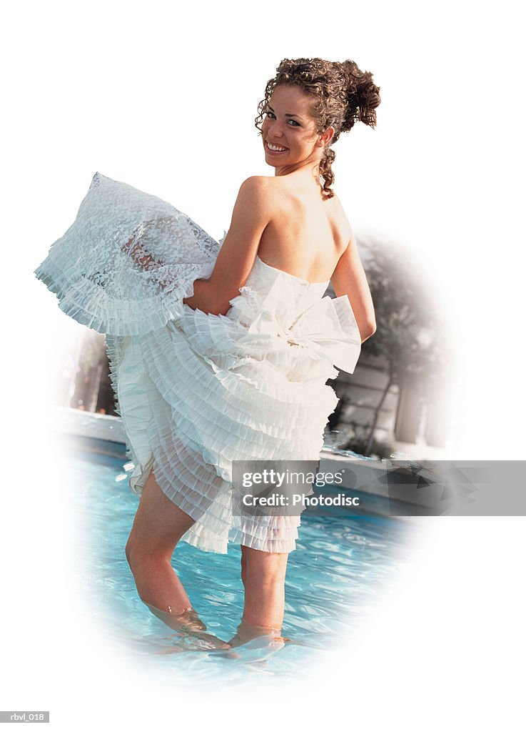 A beautiful young brunette hispanic or caucasian woman is wading in a pool as she gathers her lacey white strapless dress in her arms and smiles over her shoulder